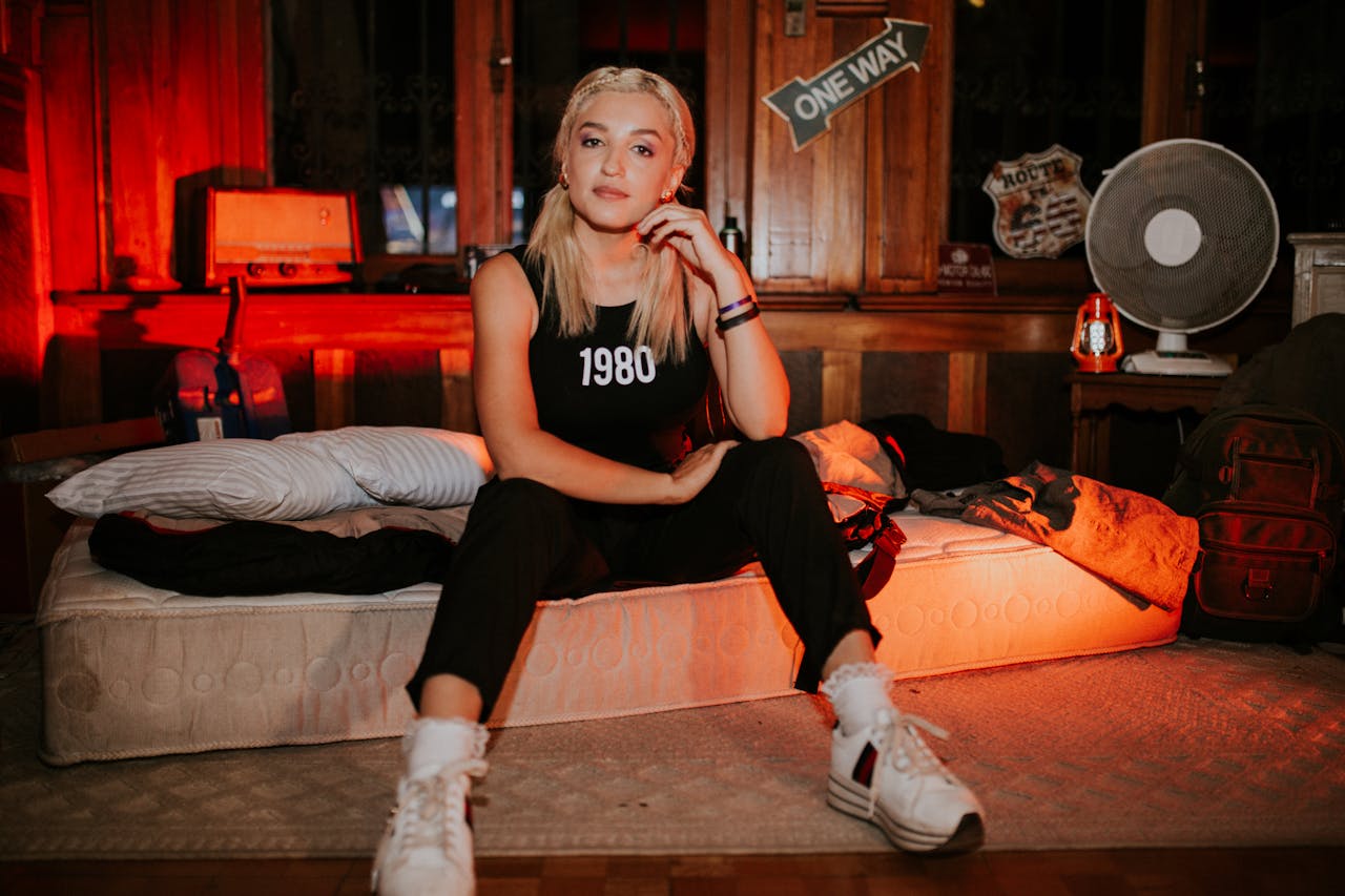 A young woman sitting on a mattress in a vintage-style bedroom with warm lighting.